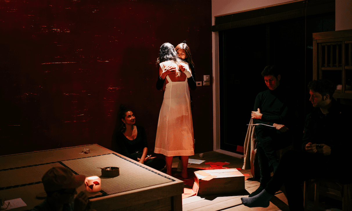 Two women embrace in a dim room while others sit and watch, during a performance